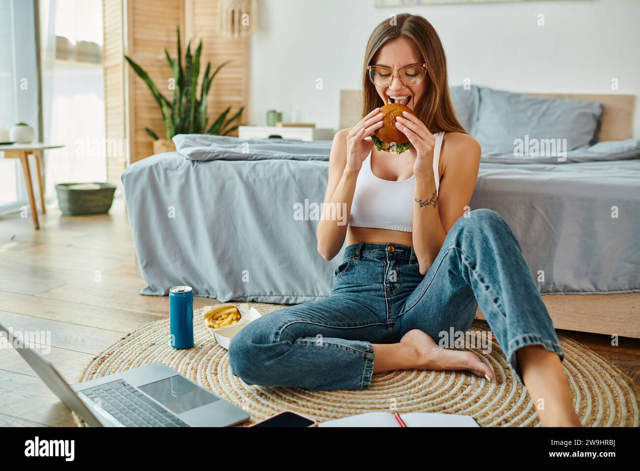 donna allegra e attraente in un abbigliamento informale seduta sul pavimento mentre lavora e si gusta un hamburger Foto Stock