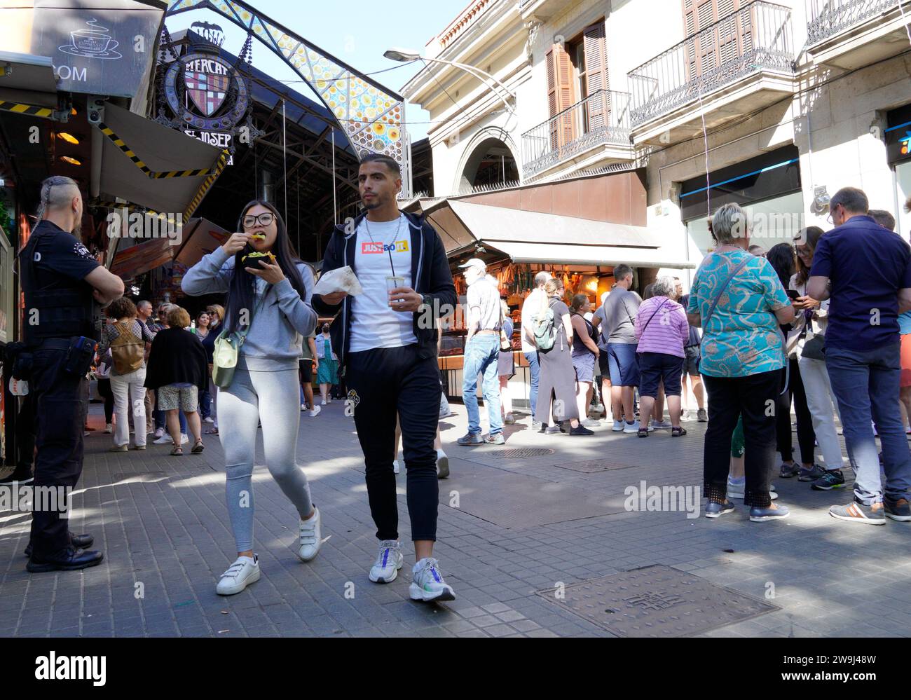 Barcellona, Spagna - 26 maggio 2022: Due di diverse origini etniche vestite di abbigliamento sportivo mangiano mentre escono dal mercato la Boqueria, un luogo tipico Foto Stock