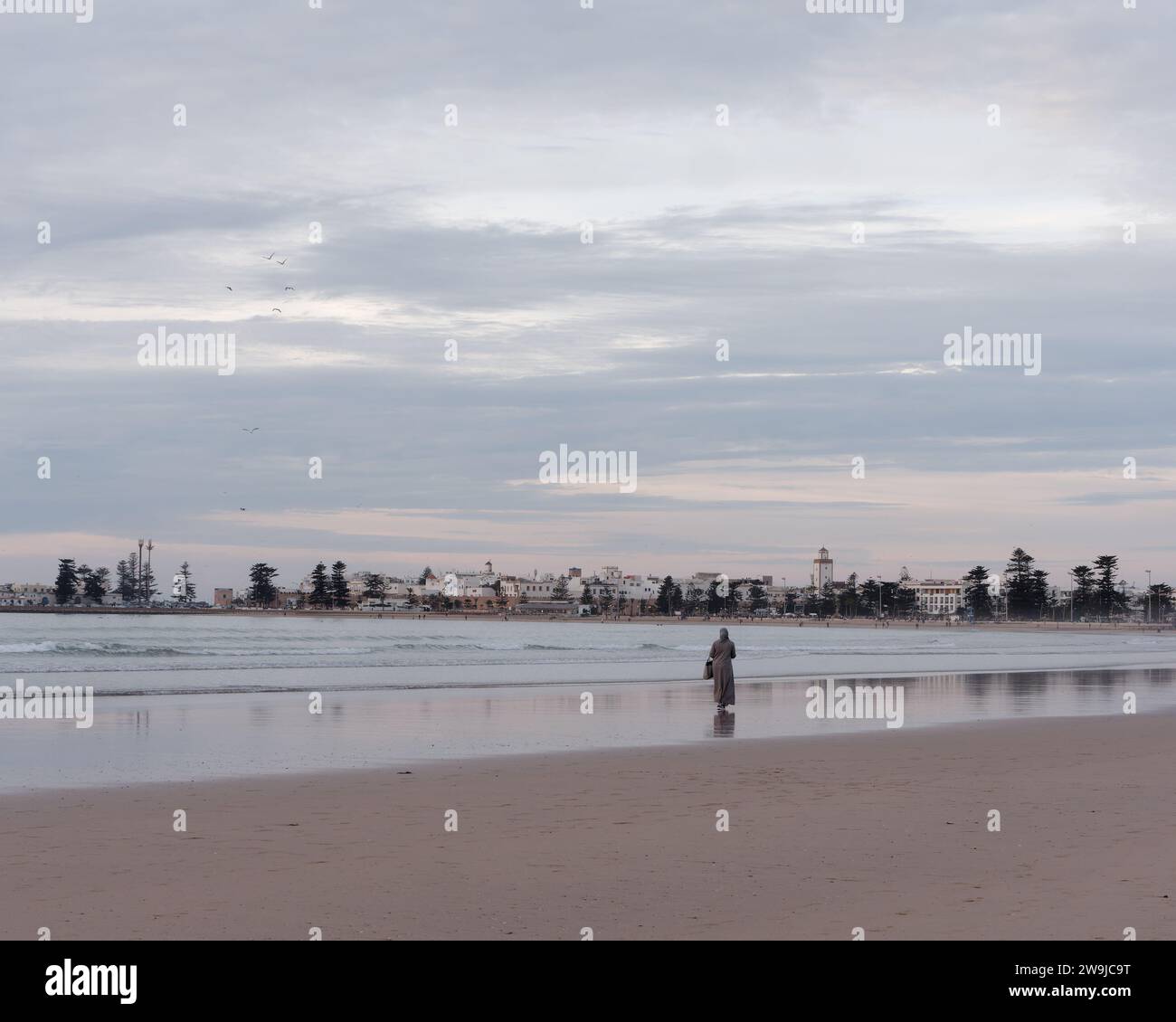 Una donna con abiti tradizionali si erge sulla spiaggia in una serata d'inverno con la storica Medina alle spalle a Essaouira, in Marocco. 28 dicembre 2023 Foto Stock