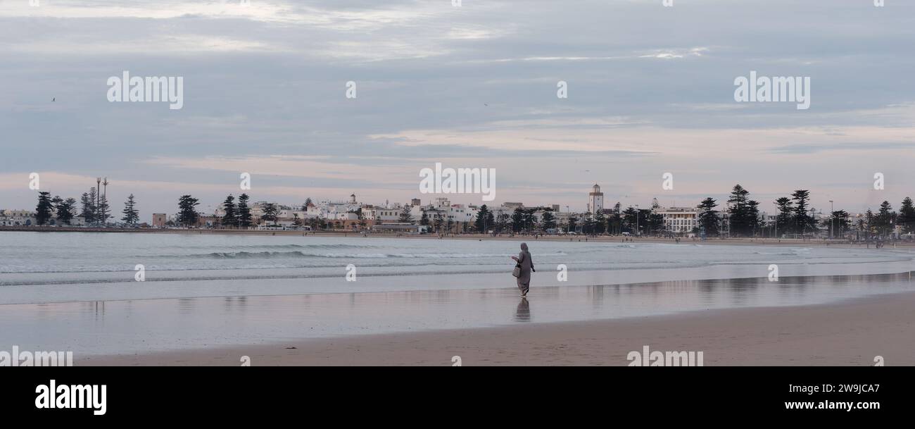 Una donna con abiti tradizionali si erge sulla spiaggia in una serata d'inverno con la storica Medina alle spalle a Essaouira, in Marocco. 28 dicembre 2023 Foto Stock