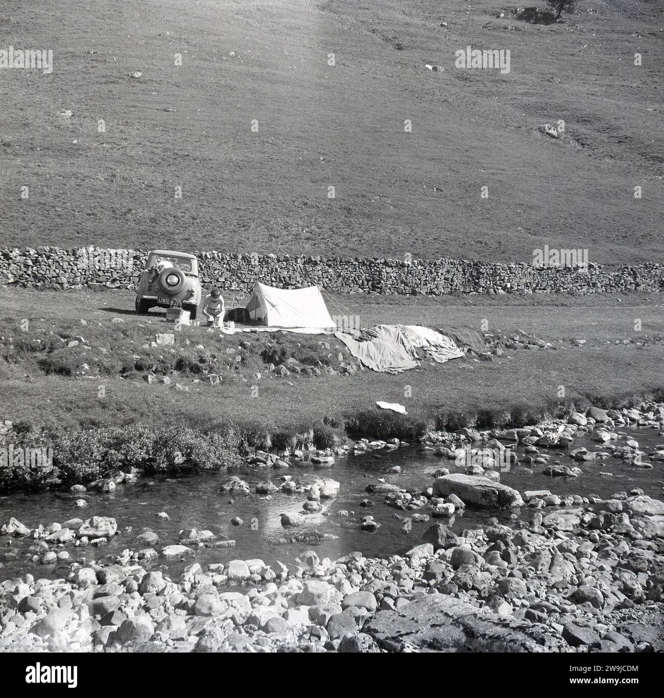 Anni '1960, storico, estivo e campeggio presso un ruscello rurale su un pezzo di terra pianeggiante vicino a un muro di pietra nel Hilllly Peak District, Inghilterra, Regno Unito. Auto dell'epoca con targa UWR 475. Foto Stock