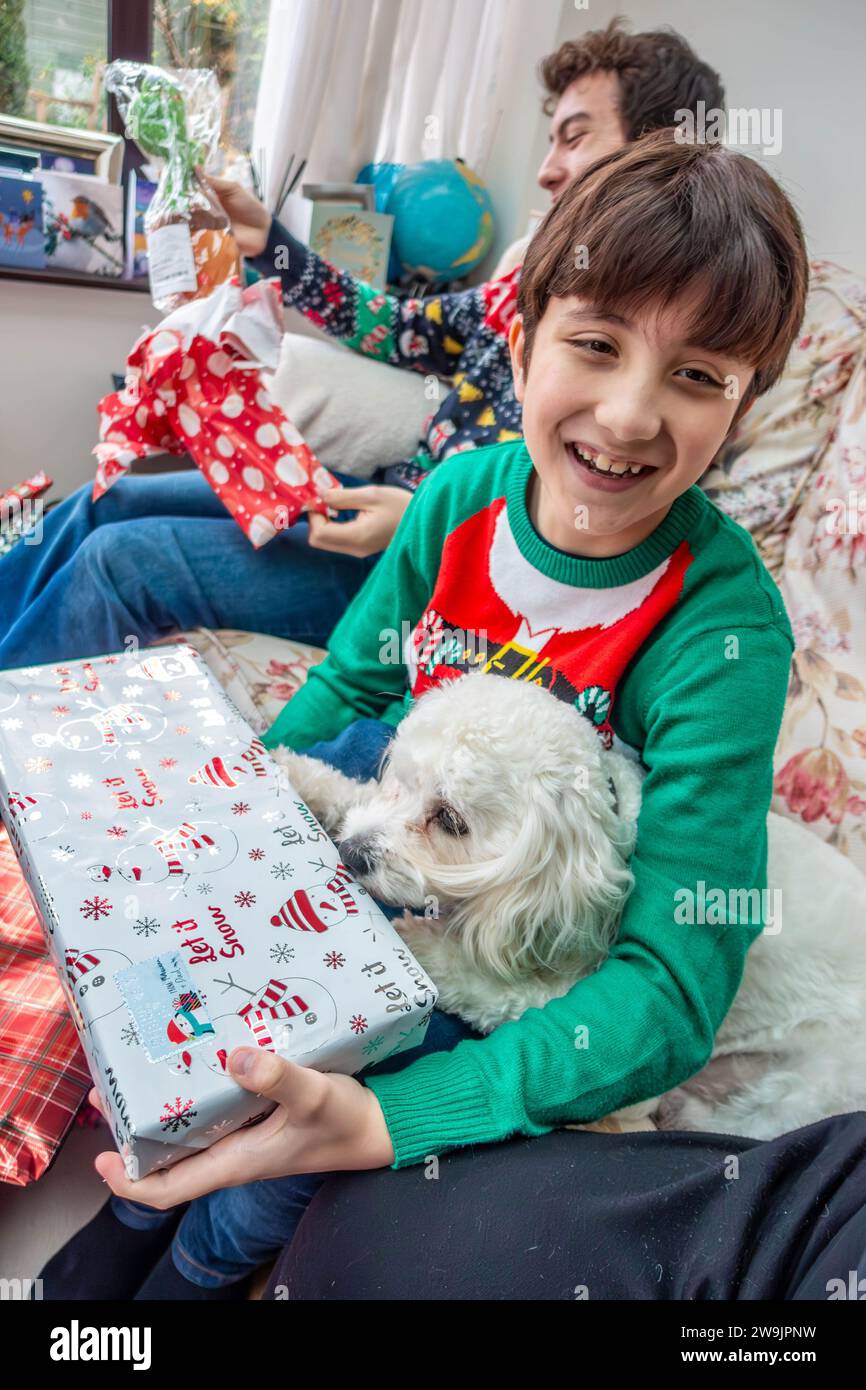 Un ragazzo che indossa un maglione di Natale tiene un regalo di Natale mentre coccola un piccolo cane cavapoo bianco. Foto Stock