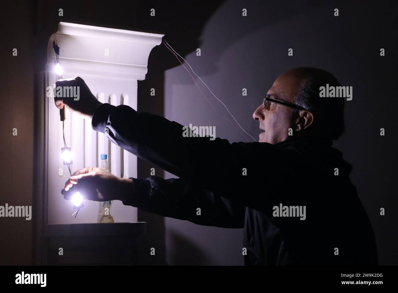 Al Hasakah. 28 dicembre 2023. Ghassan al-Hamoud, un insegnante di 51 anni, collega una piccola luce a una batteria a casa nella provincia di al-Hasakah, nel nord-est della Siria, il 28 dicembre 2023. Nel cuore della città di al-Hasakah nel nord-est della Siria, una famiglia di sei lotte sotto il peso delle sfide imposte dalla presenza delle forze statunitensi nel nord-est e nella Siria orientale. Crediti: Str/Xinhua/Alamy Live News Foto Stock