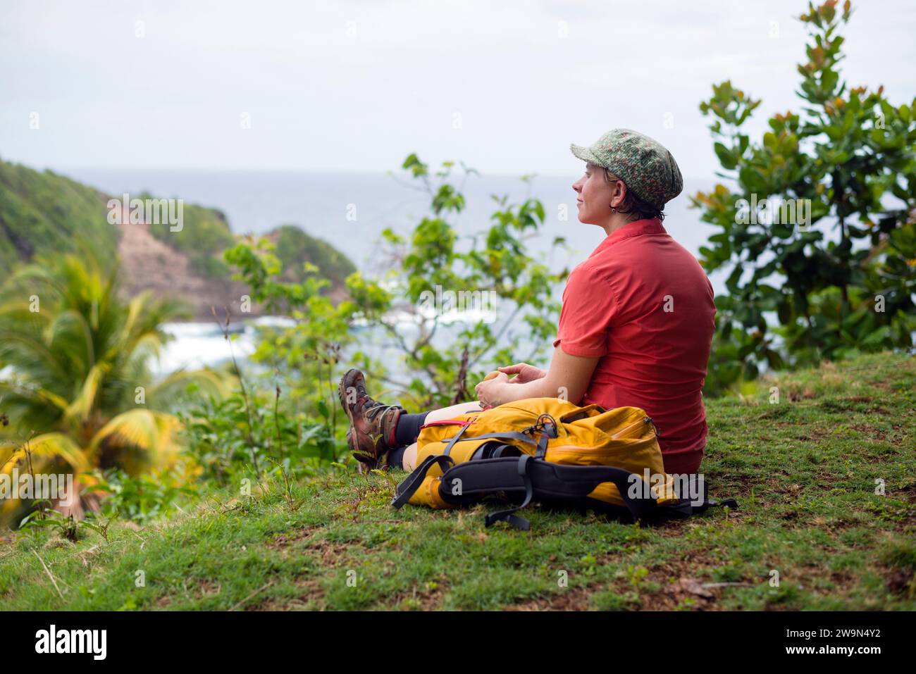 Un viaggiatore con zaino in spalla fa una pausa affacciandosi sull'Oceano Atlantico nel territorio di Kalinago sul segmento 6 del Waitukubuli National Trail sull'isola caraibica di Dominica. Foto Stock