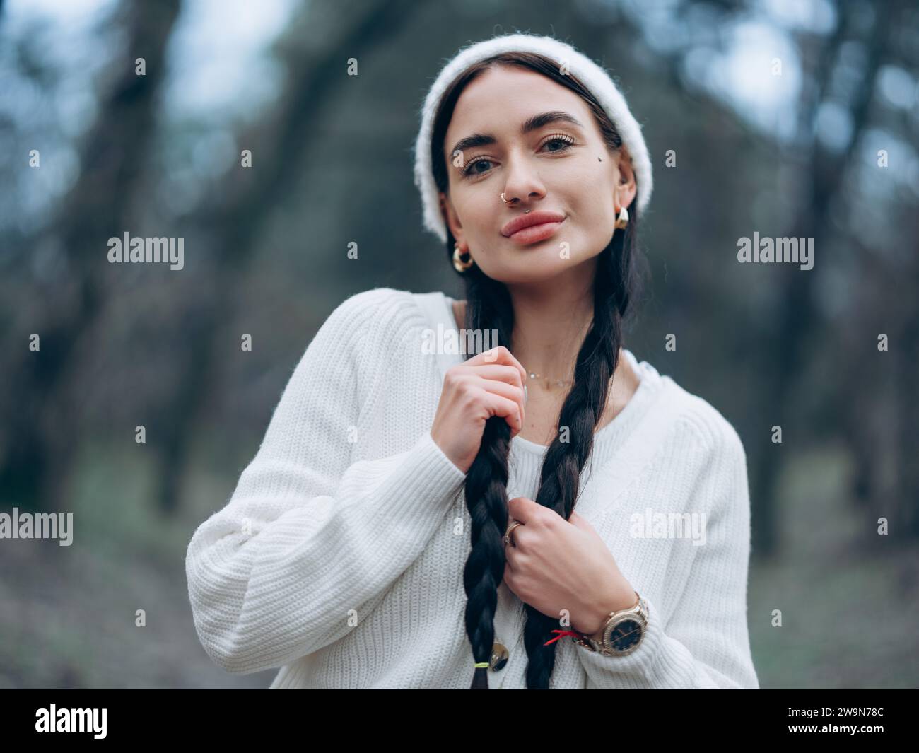 Ritratto di una giovane donna sorridente con cappello bianco e maglione con capelli intrecciati e piercing sul viso sullo sfondo della foresta. Foto Stock