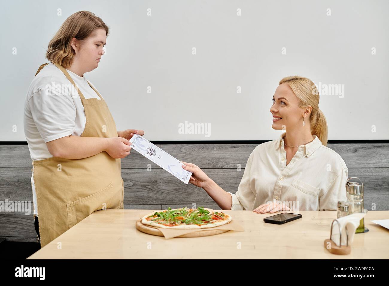 giovane cameriera con disabilità mentale che dà la carta del menu a una donna felice seduta in un moderno bar Foto Stock