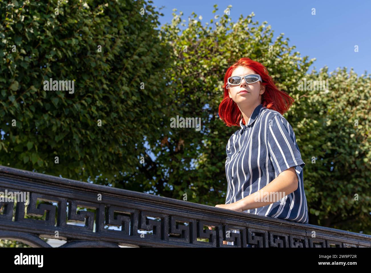 Un'adolescente alla moda con i capelli rossi e gli occhiali da sole alla moda si gode una piacevole passeggiata nel parco cittadino in una luminosa e soleggiata giornata d'estate Foto Stock