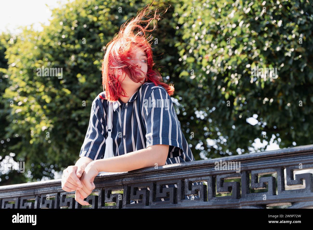 Giovane donna dai vivaci capelli rossi che cammina in un parco cittadino mentre il vento soffia tra gli alberi. Rappresenta la generazione Z demografica con il suo truffatore Foto Stock