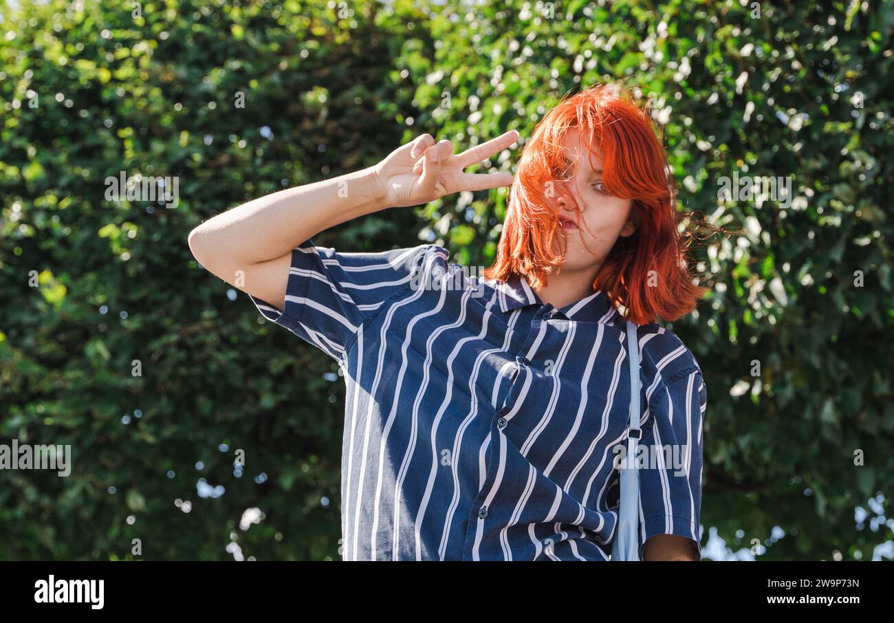 Un'allegra adolescente, rappresentativa della generazione Z, con i capelli rossi vivaci, passeggiando gioiosamente in un parco cittadino ed esprimendo un segno di vittoria con h Foto Stock