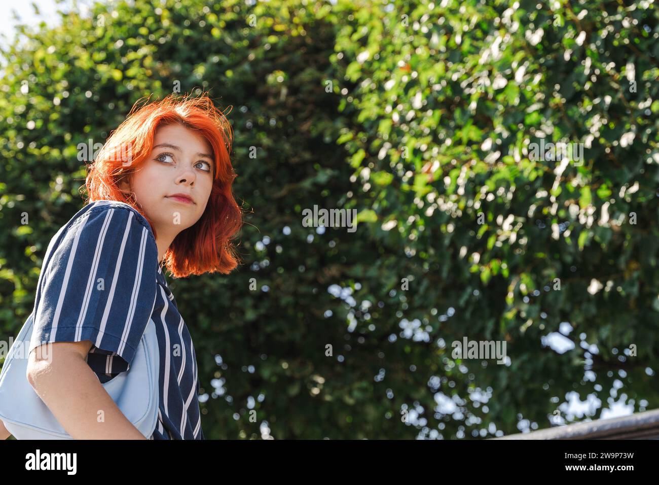 Un ritratto di una giovane ragazza adolescente della generazione Z dai capelli rossi che si gode una piacevole passeggiata in un bellissimo parco cittadino in una soleggiata giornata estiva, circondata da una lussureggiante vegetazione Foto Stock