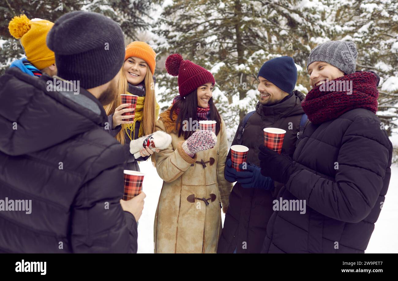 Felice ed eccitante gruppo di amici in piedi nella foresta bevendo tè caldo e chiacchierando durante la passeggiata invernale. Foto Stock