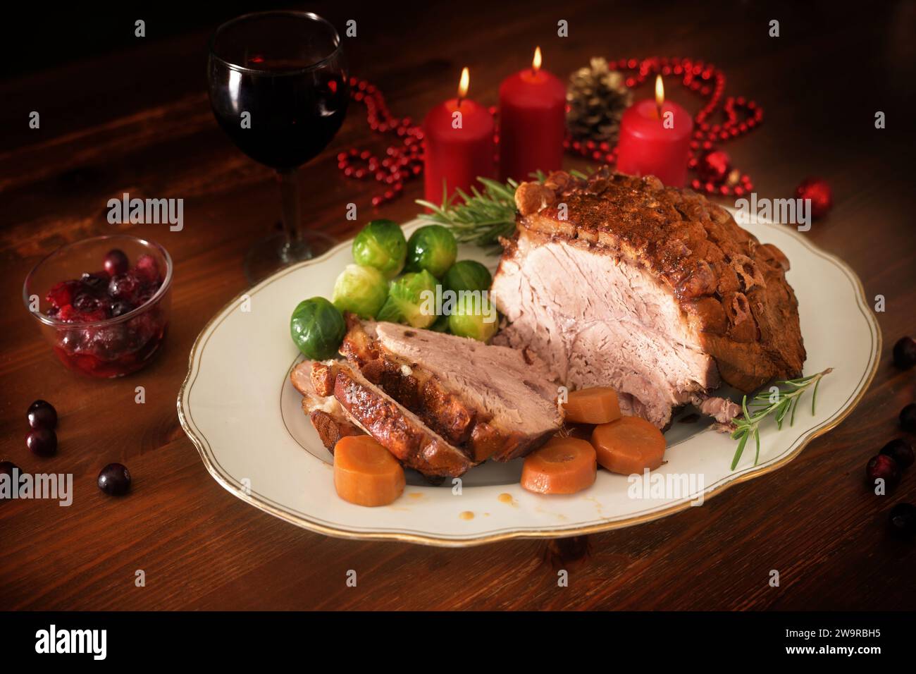 Arrosto di maiale o prosciutto di Natale con crosta, servito con verdure e mirtilli rossi su un tavolo di legno scuro con candele, spazio per la copia, fuoco selezionato, d stretto Foto Stock