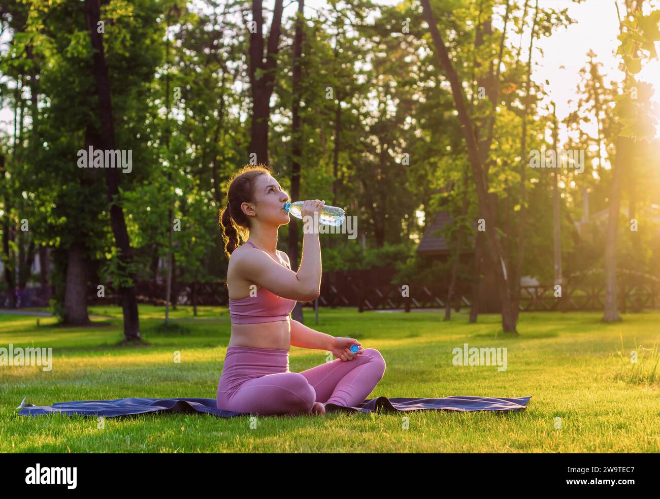 La giovane ragazza beve acqua mentre fa yoga all'aperto nel parco al tramonto. Concetto di stile di vita sano. Foto Stock