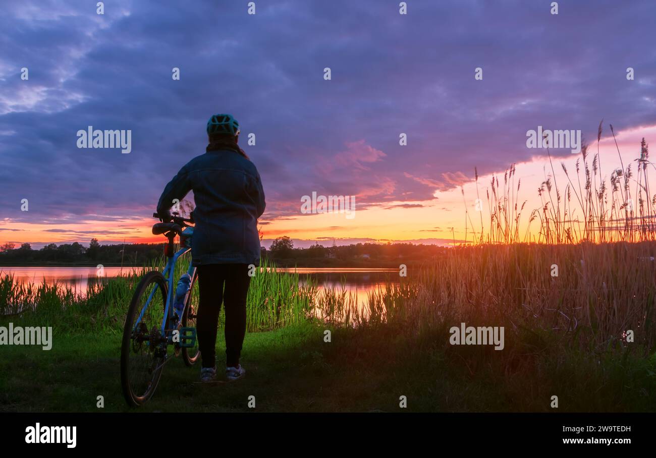 Una ragazza con una bici da strada si trova sulla riva di un lago contro un bel cielo al tramonto. Il concetto di uno stile di vita attivo. Foto Stock