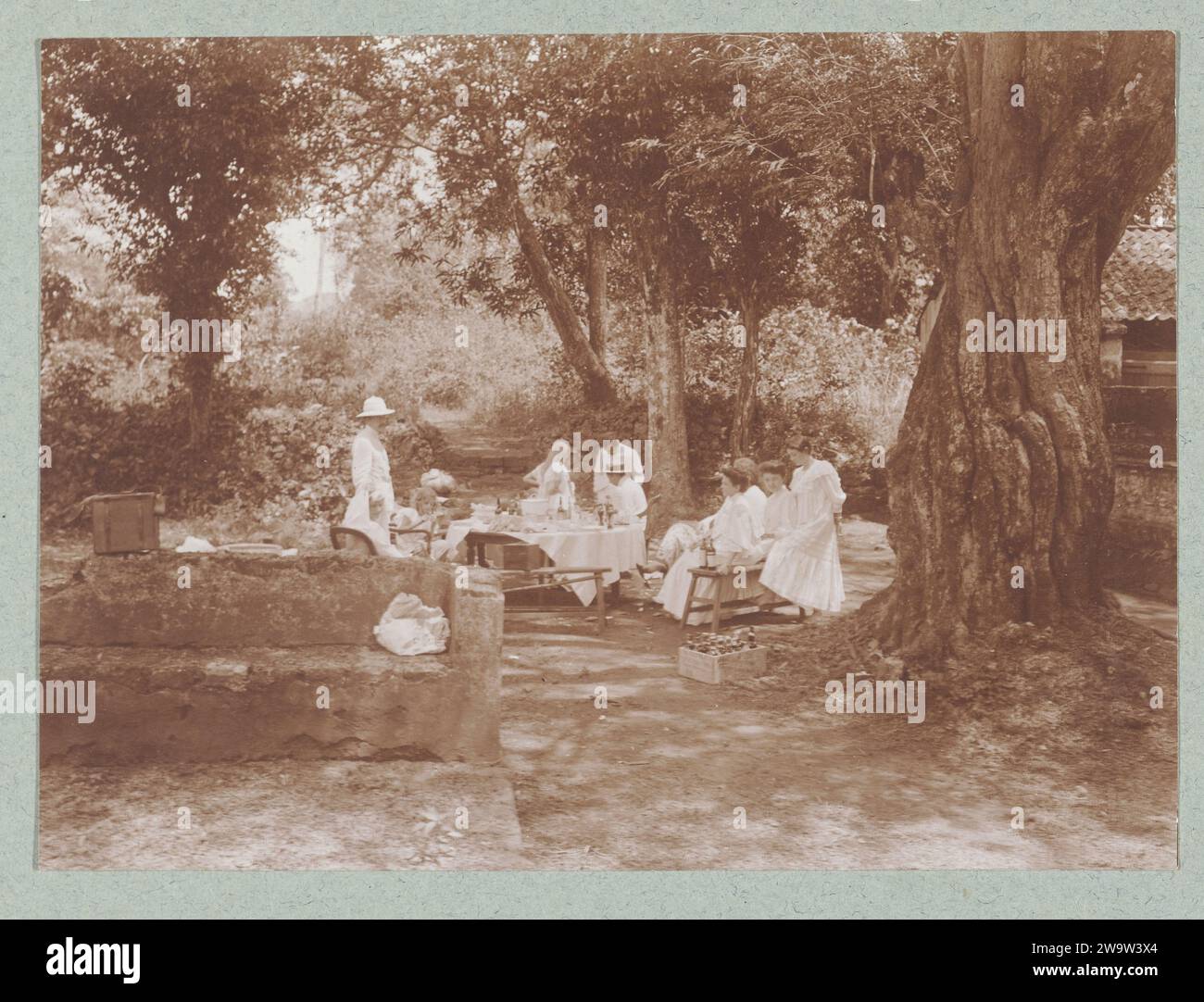 Un tavolo fisso ai tropici con gente europea vestita di bianco., c. 1910 fotografa gli uomini che indossano uniformi tropicali bianche di cui un uomo con un casco tropicale, altri due uomini indossano un cappello di paglia, le donne indossano abiti bianchi o gonne batik con hes bianche. Foto dall'album "Photographies". Madura (possibilmente)Olanda (possibilmente) cartone. carta Foto Stock