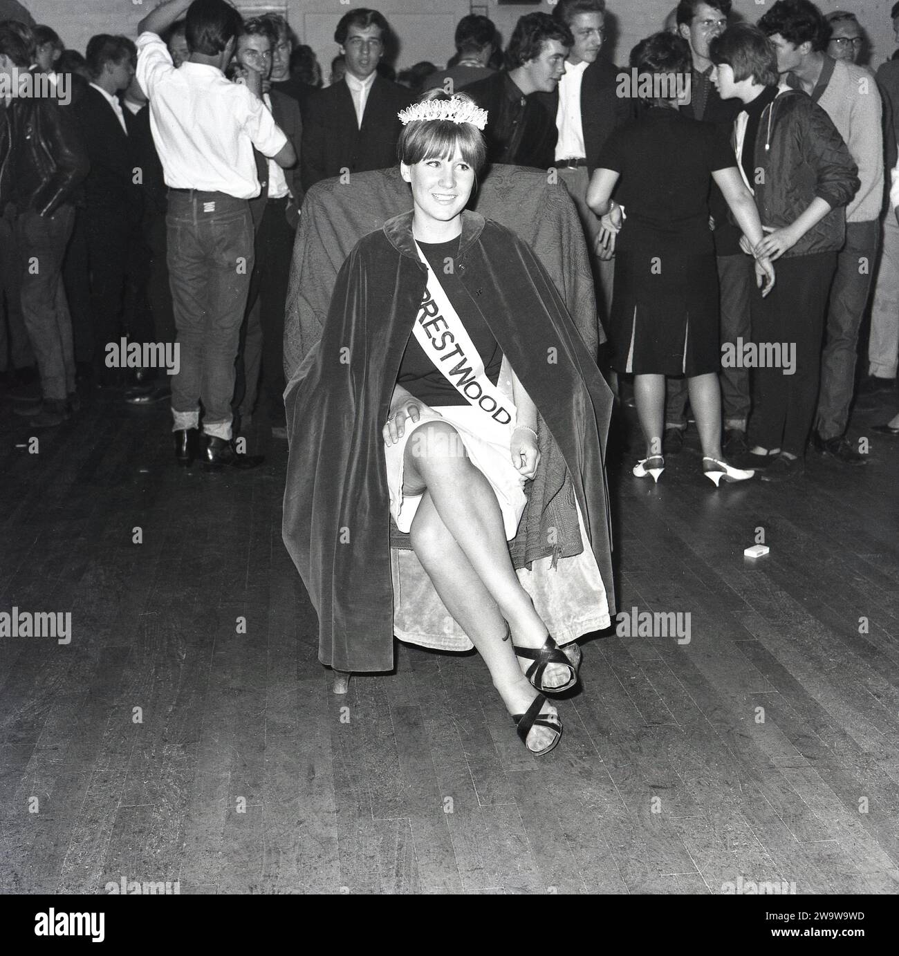 1964, storica, bella regina.... Al centro di un municipio con altri giovani presenti, una giovane donna, con corona, fascia e mantello seduto su una sedia dopo essere stata incoronata come "Miss Prestwood". L'antico villaggio di Prestwood nelle Chilterns, vicino a Great Missenden nel Buckinghamshire, Inghilterra, risale all'epoca anglosassone. Foto Stock