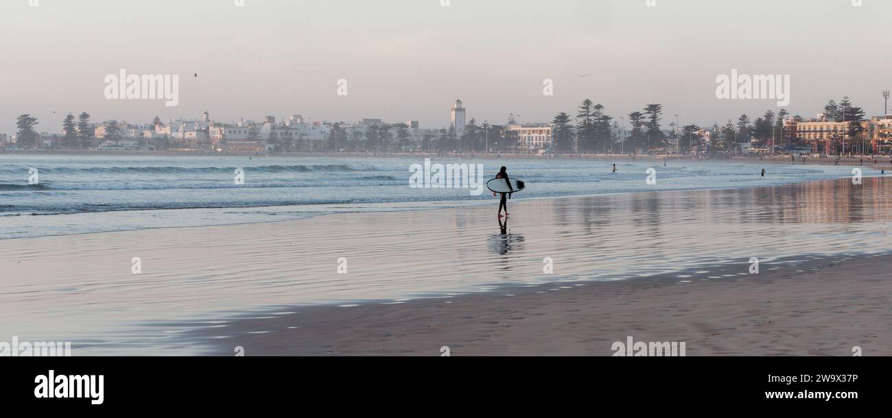 Il surfista solitario esce dall'acqua su una spiaggia sabbiosa avvicinandosi al tramonto con la Medina alle spalle nella città di Essaouira, in Marocco. 30 dicembre 2023 Foto Stock