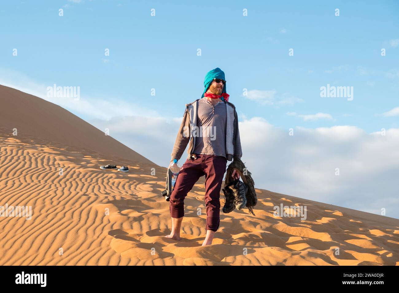 Il turista maschile con il copricapo di sciarpa si trova a piedi nudi su una duna di sabbia nel mare di Erg Cebbi, nel deserto del Sahara, nell'Africa settentrionale Foto Stock