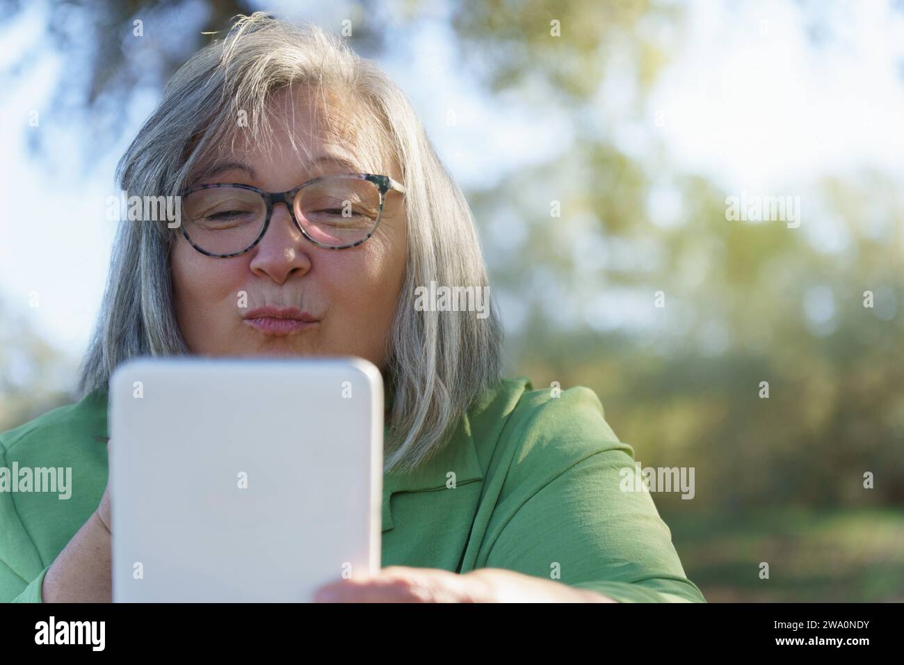 Donna matura dai capelli bianchi con occhiali che si bacia sul suo tablet digitale durante una videochiamata in campagna Foto Stock