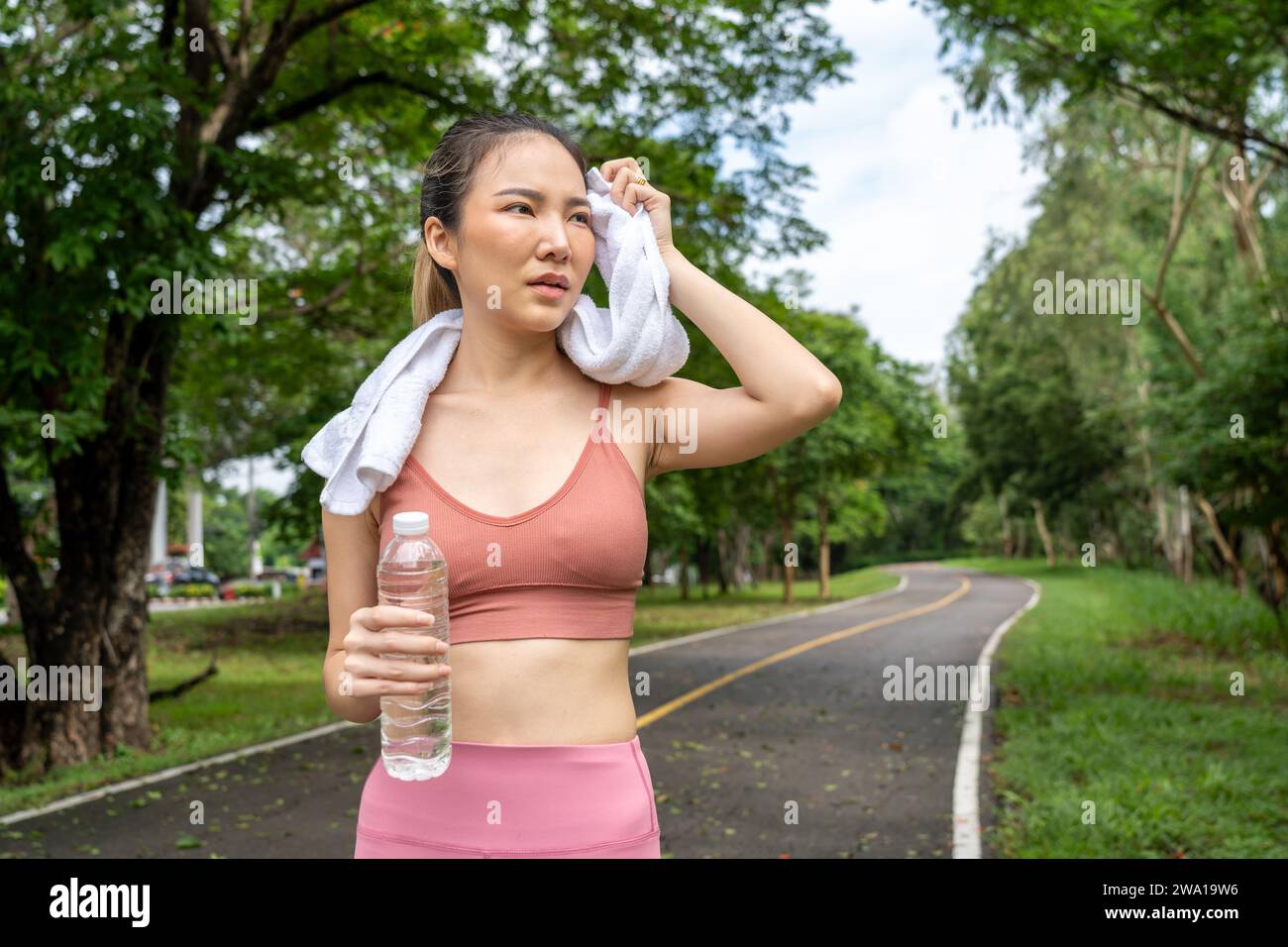 Giovane attraente donna asiatica che si toglie il sudore dalla fronte durante la pausa in acqua dalla corsa di esercizio mattutina su una pista di corsa di un locale Foto Stock