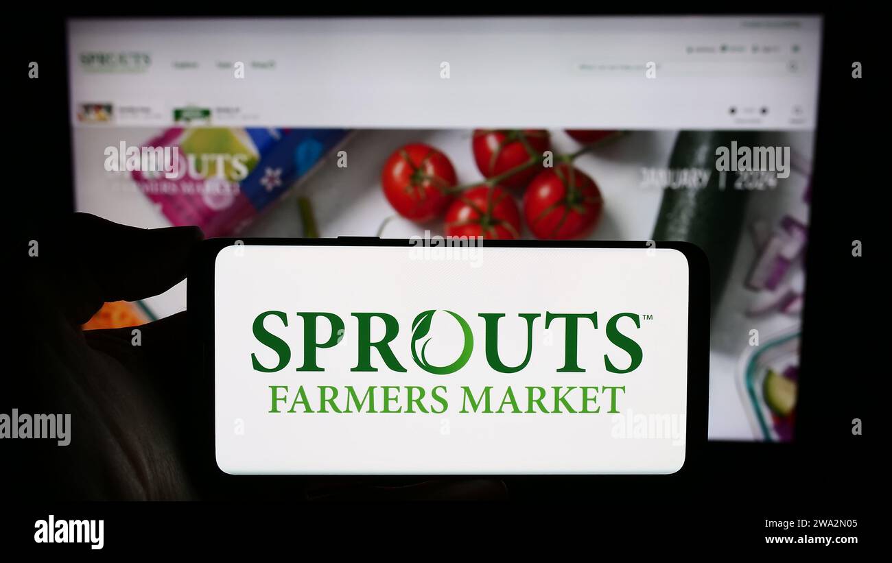 Persona che possiede uno smartphone con il logo della società di vendita al dettaglio statunitense sprouts Farmers Market Inc. Davanti al sito Web. Concentrarsi sul display del telefono. Foto Stock