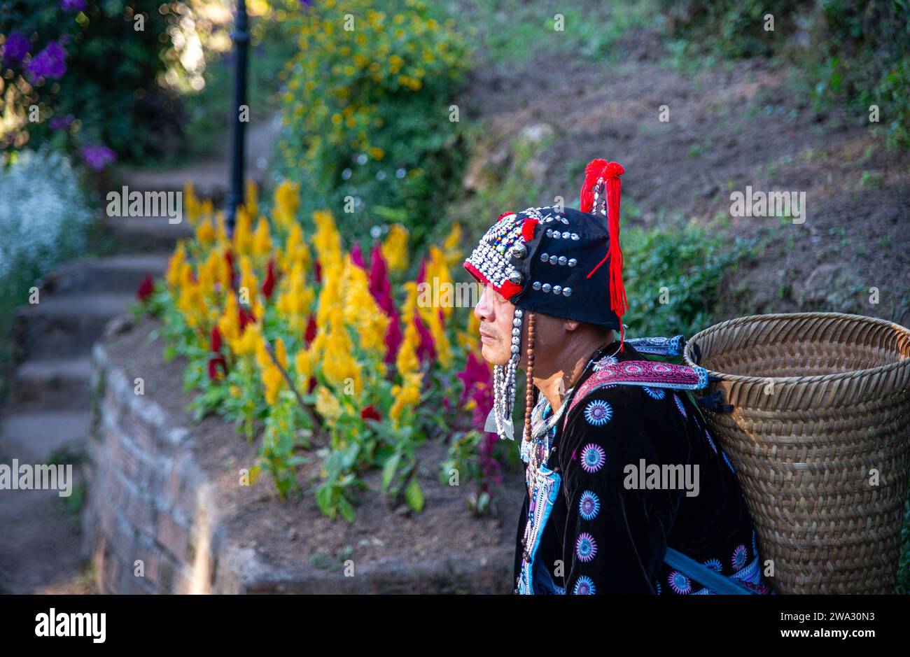 20 febbraio-2023-Chiang Rai- Thailandia- Vecchio con cesto sulla schiena e vestito con costumi tradizionali. Sullo sfondo ci sono fiori colorati Foto Stock