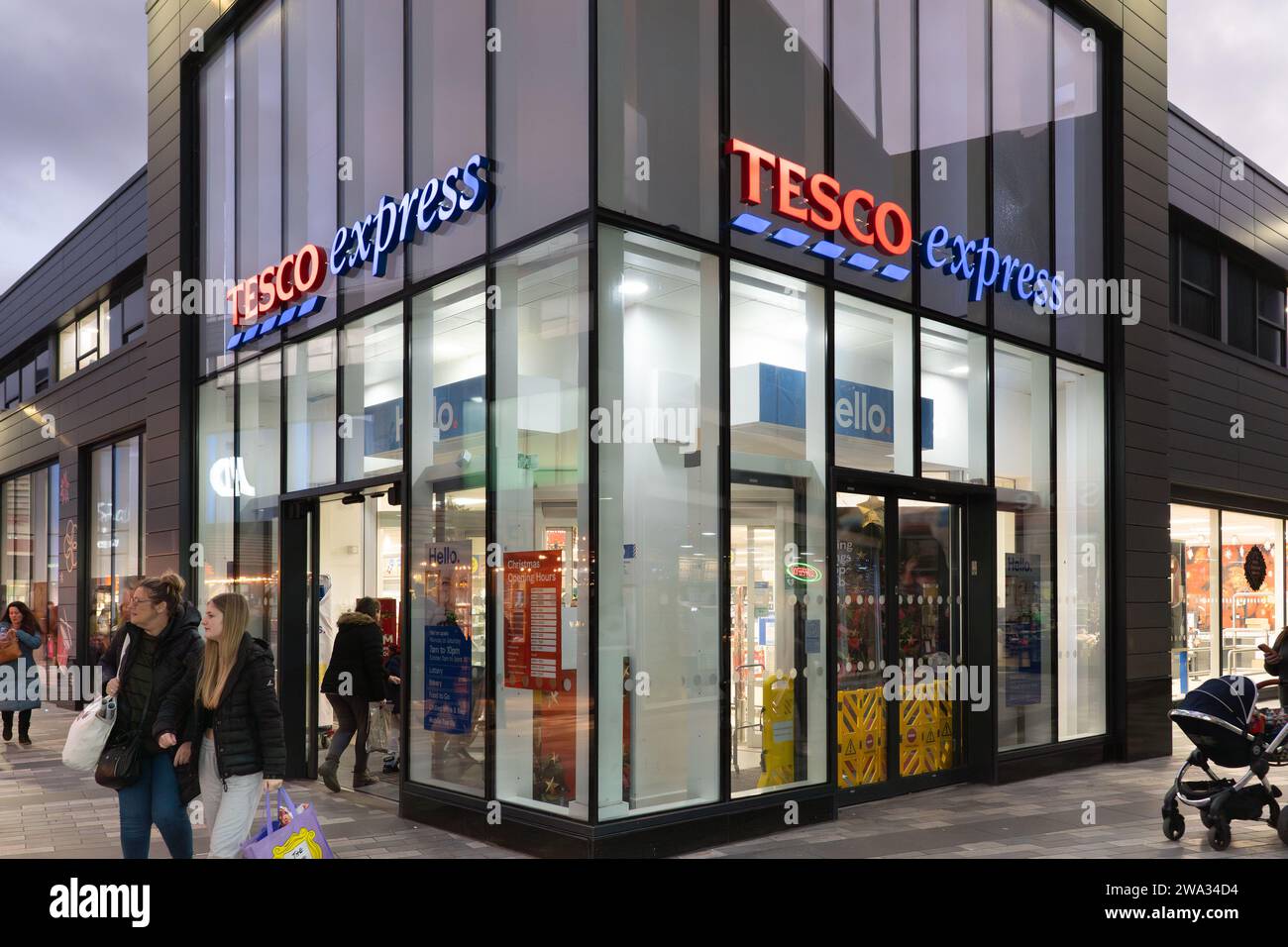 Gli acquirenti entrano e escono da un Tesco Express al crepuscolo nei centri commerciali di Basingstoke, Regno Unito. Tema: Costo della vita, inflazione alimentare, costo dello shopping Foto Stock