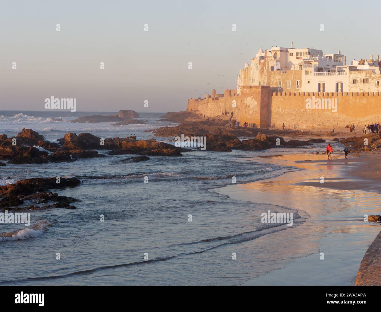 La gente cammina su una spiaggia rocciosa e sabbiosa durante un tramonto invernale con le mura della città e la medina alle spalle a Essaouira, Marocco, 1 gennaio 2024 Foto Stock
