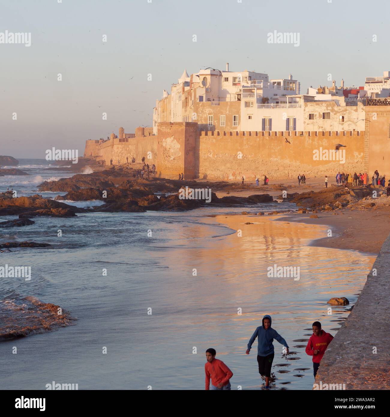 Le persone corrono lungo la spiaggia con la Medina e le mura della città alle spalle durante un tramonto invernale nella città di Essaouira, Marocco, il 1° gennaio 2024 Foto Stock