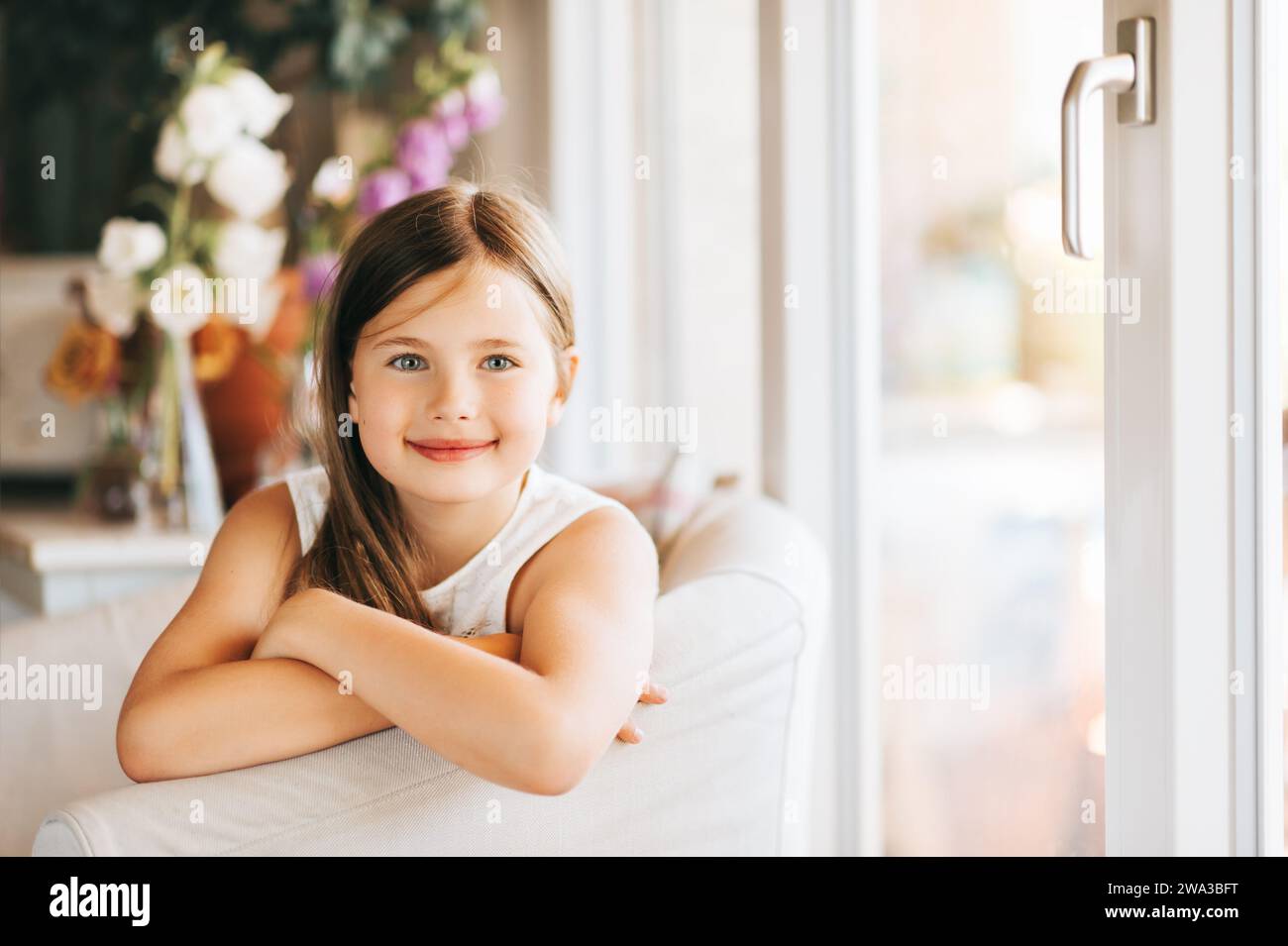 Una bambina carina seduta su una sedia bianca accanto alla finestra di casa Foto Stock