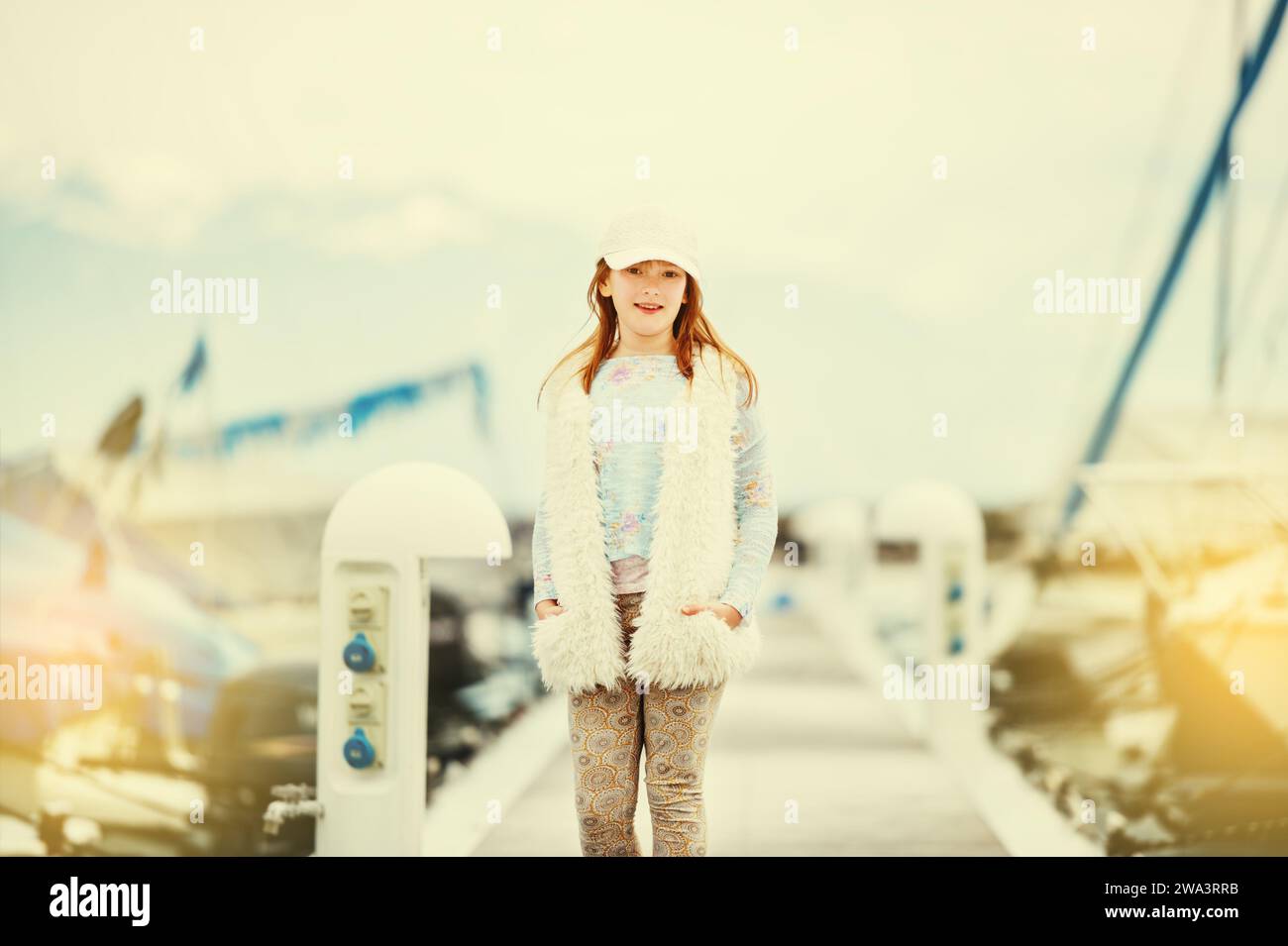Ritratto di moda all'aperto di una giovane ragazza preadolescente che indossa un berretto bianco, un gilet in pelliccia sintetica, che gioca in un porto in una giornata molto soleggiata e con un'immagine tonica Foto Stock