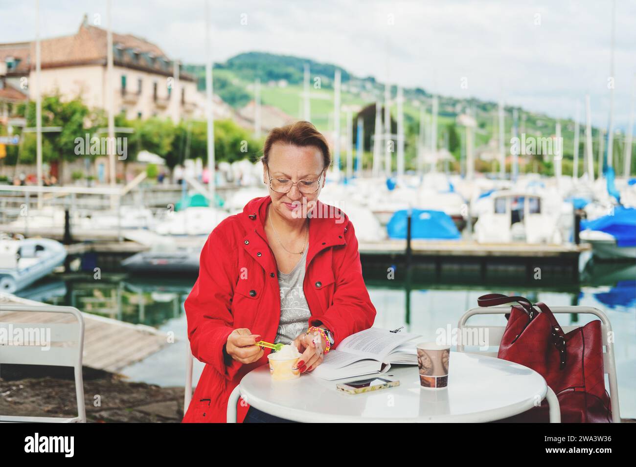 Donna di 50-60 anni che riposa all'aperto, leggendo un libro in una caffetteria sul lago di Ginevra, Svizzera Foto Stock