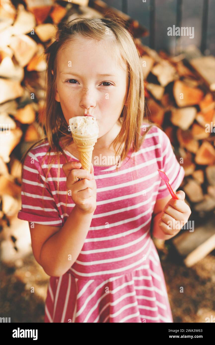 Graziosa bambina di 6 anni che mangia gelati all'aperto, indossa un abito a strisce rosa, piaceri estivi per i bambini Foto Stock