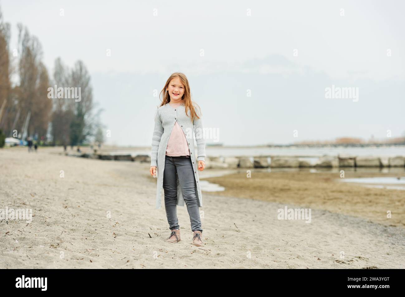 Adorabile bambina di 8-9 anni che gioca vicino al lago, indossa pantaloni grigi e un lungo cardigan Foto Stock