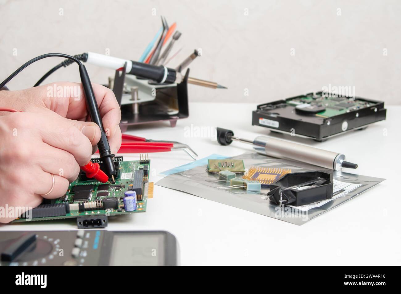 Primo piano delle mani di un tecnico in un'officina. Il riparatore utilizza un multimetro per misurare i parametri di un dispositivo elettronico su un tavolo. Foto Stock