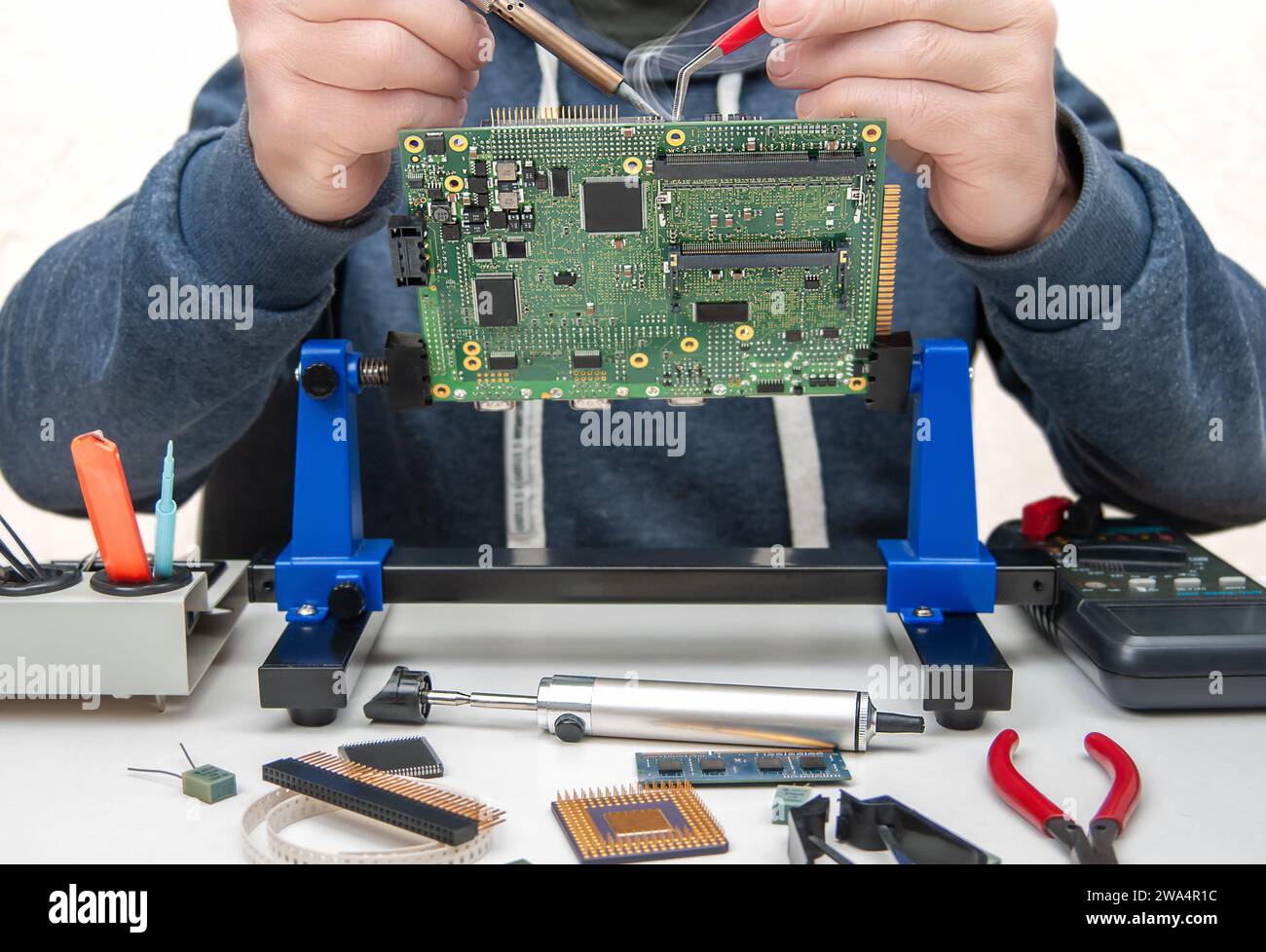 Primo piano delle mani di un tecnico in un'officina. Il riparatore è un circuito stampato di saldatura di un dispositivo elettronico sul tavolo con pinzette. Foto Stock