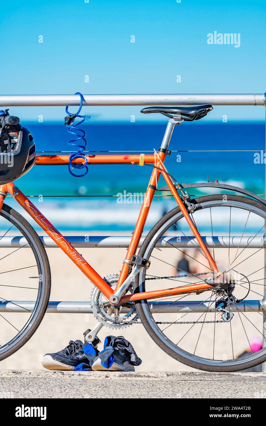 Una bicicletta da strada e un casco da bicicletta contro una ringhiera d'acciaio e un paio di corridori con Bondi Beach a Sydney, Australia, sfocati sullo sfondo Foto Stock