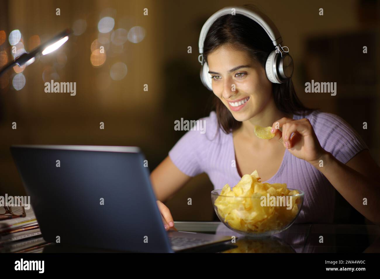 Donna felice che indossa le cuffie di notte mentre guarda un video su un computer portatile mentre mangia patatine a casa Foto Stock