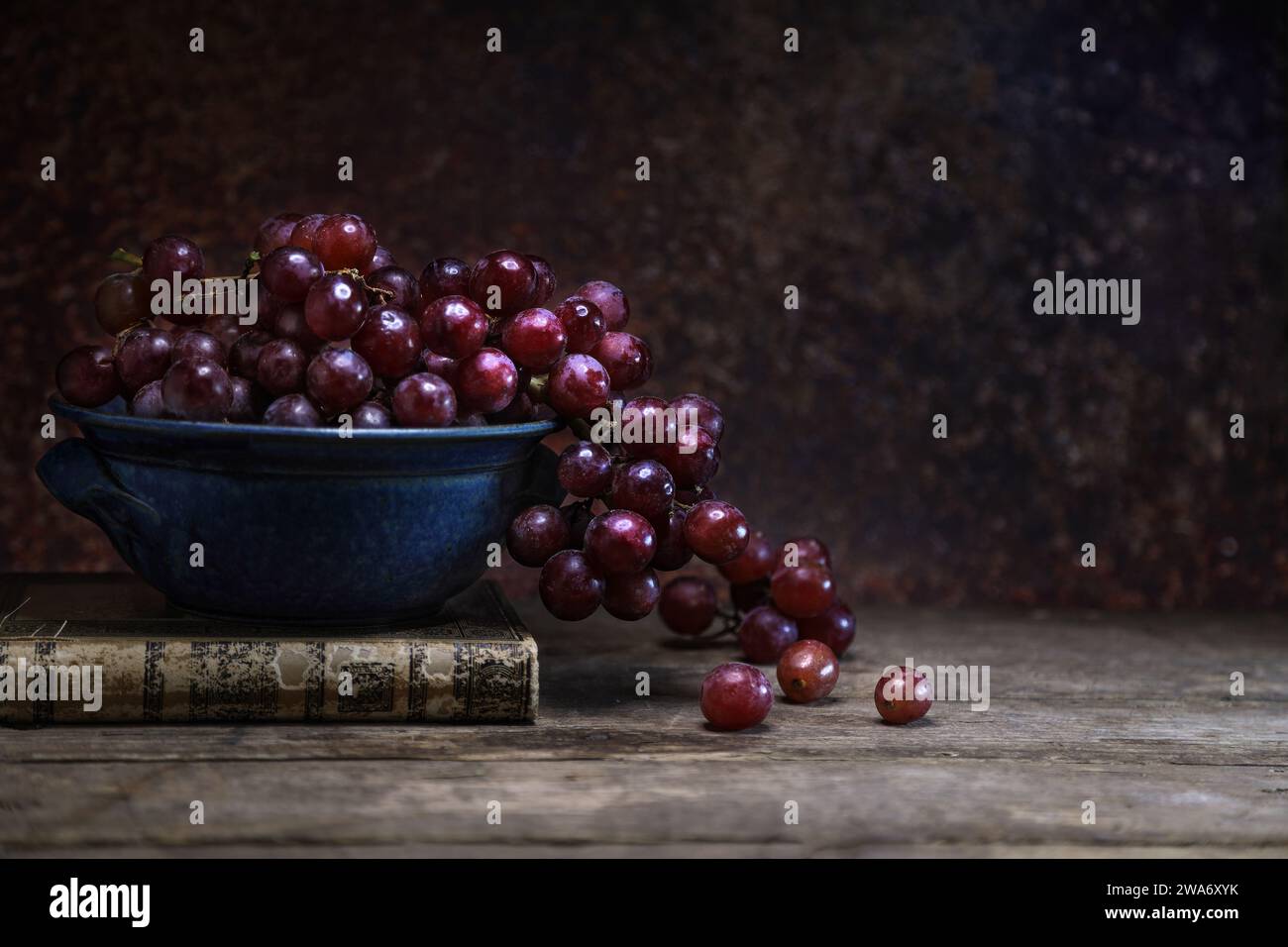 Uva rossa fresca in una ciotola blu con un libro d'epoca su tavola e sfondo di legno - scuro e lunoso Foto Stock