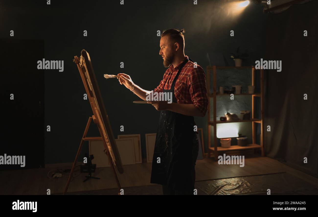 Artista maschile di talento che utilizza pennello crea un capolavoro. Studio creativo scuro con ampia tela Foto Stock