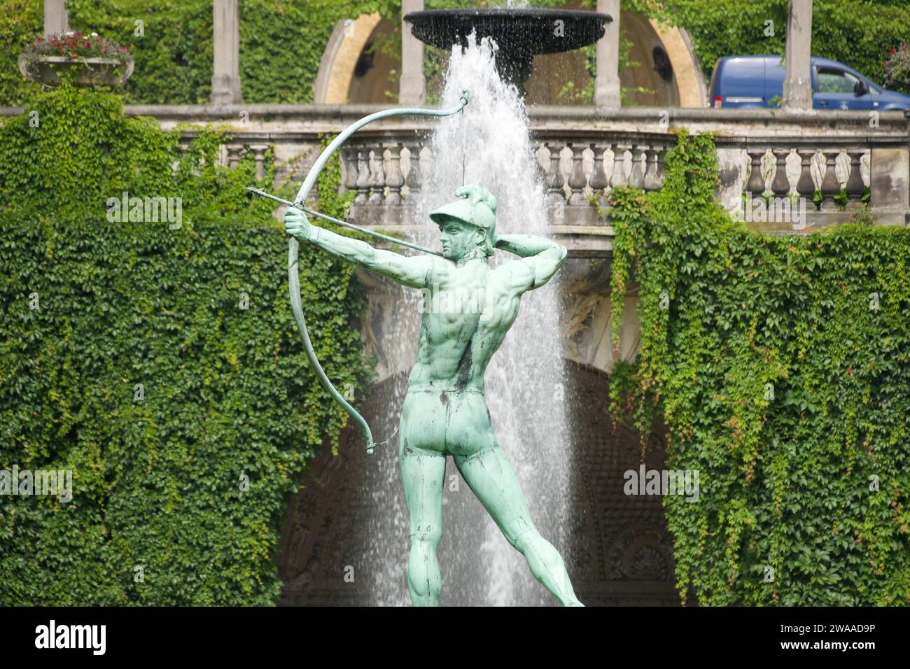 Questa vivace scultura di un arciere, adagiata su uno sfondo di vegetazione lussureggiante e di una fontana che scorre, racchiude lo spirito vibrante dell'atrio pubblico di Potsdam Foto Stock