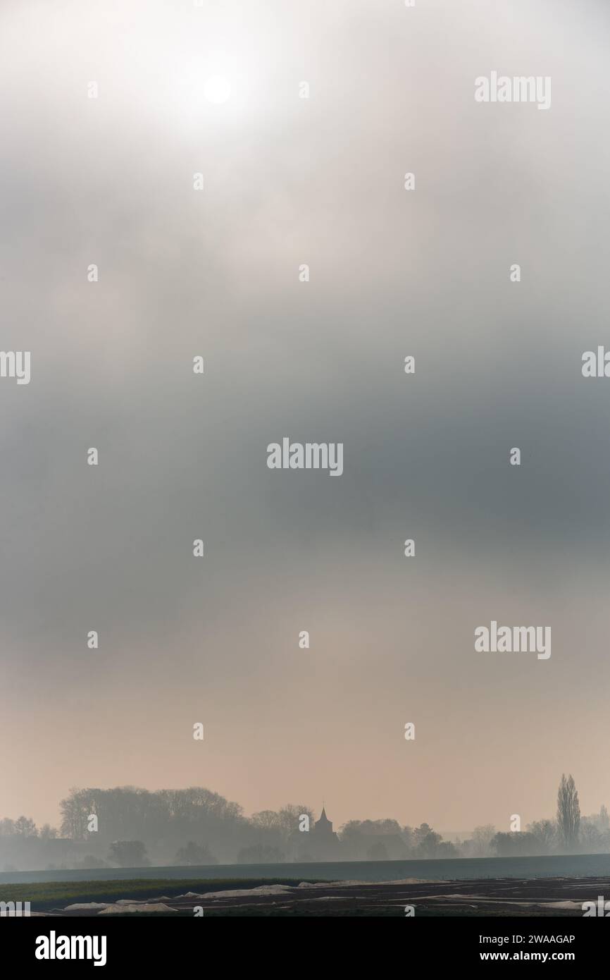 In un cielo pieno di nebbia, il sole splende debolmente sulla campagna. Campanile all'orizzonte. Foto Stock