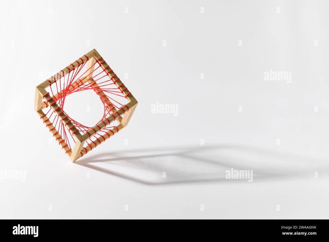 Visualizzazione brillante di cubo in legno massello platonico instabile con filo in tessuto rosso in piedi sul bordo su sfondo bianco brillante con ombra Foto Stock