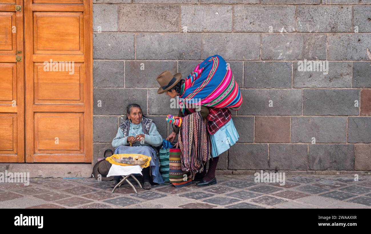 Cusco, Perù - 24 ottobre 2023: Donne peruviane che vendono souvenir e cibo per i turisti seduti di fronte a un muro di pietra vicino alla strada Foto Stock