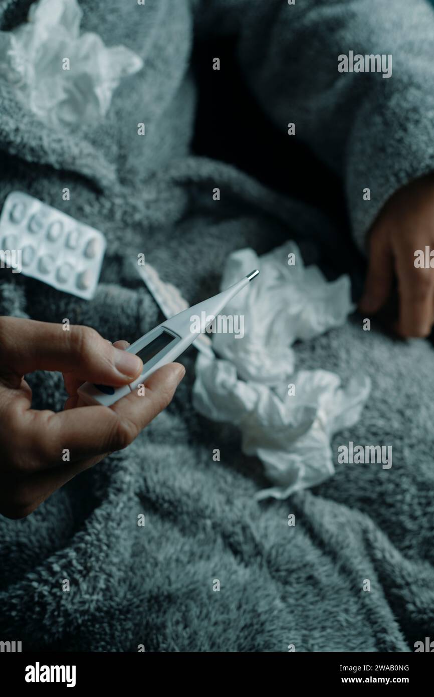 un uomo malato a casa, che indossa un soffice abito da casa grigio, circondato da tessuti usati, sta per misurare la sua temperatura con un termometro Foto Stock
