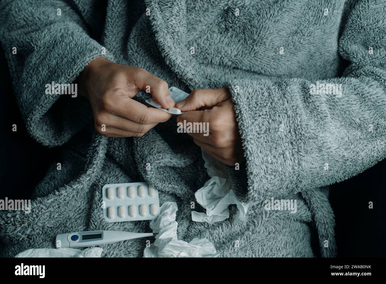 primo piano di un uomo malato a casa, che indossa un caldo abito grigio, circondato da tessuti usati, sta per prendere una pillola Foto Stock