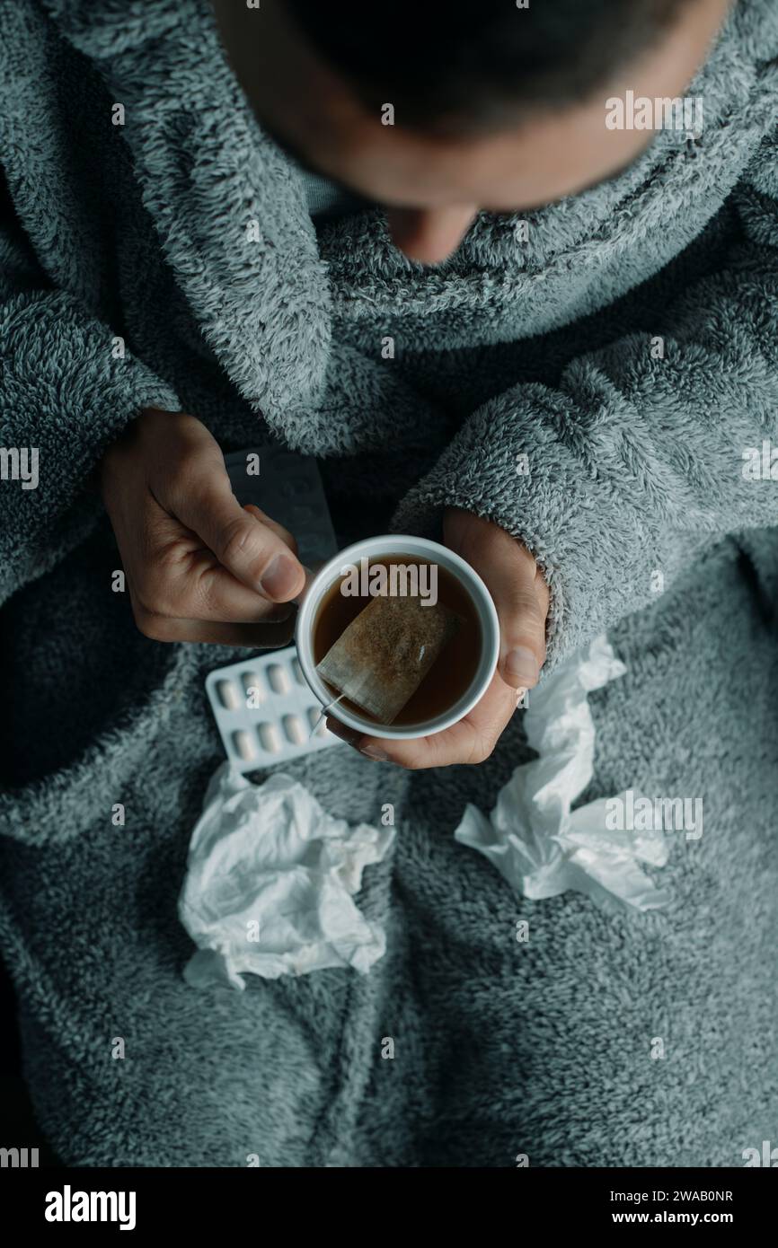 vista ad alto angolo di un uomo malato a casa, che indossa un soffice abito grigio della casa, riscaldandosi con una tazza di tè caldo alle erbe Foto Stock