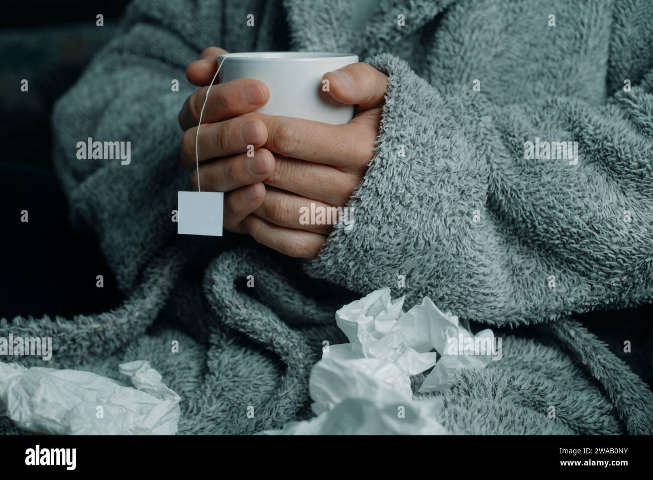un uomo malato a casa, che indossa un soffice abito da casa grigio, circondato da tessuti usati, si riscalda con una tazza di tè caldo alle erbe Foto Stock