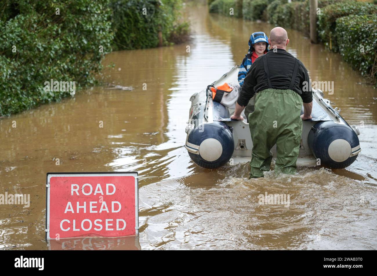 Worcester, 3 gennaio 2024 - Jack, di 7 anni e mezzo, viene trasportato in barca alla sua casa nel borgo inglese di Severn Stoke, completamente tagliato dalle inondazioni dopo che il fiume Severn ha fatto scoppiare le sue rive a causa delle continue piogge della tempesta Henk. Il pub Rose and Crown potrebbe essere visto pompare acqua mentre combatteva per rimanere asciutto con le loro difese fatte in casa contro le inondazioni. - Credito: Stop Press Media/Alamy Live News Foto Stock