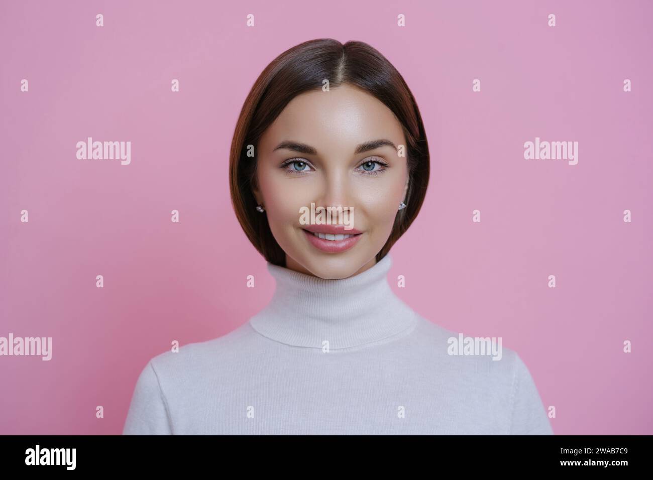 Donna elegante con capelli lucidi in un maglione dolcevita che regala un sorriso delicato sullo sfondo rosa Foto Stock